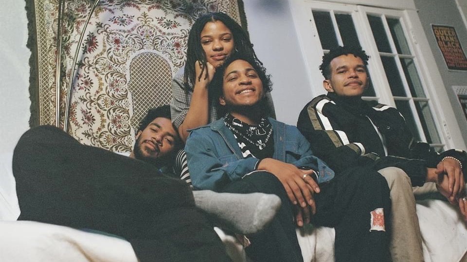 A promotional image of the band Soul Flower. The four band members sit together on couch in front of a decorative tapestry