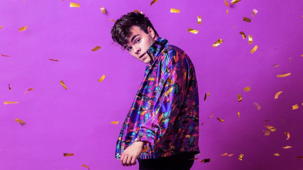 Grayson Dewolfe stands in front of a fuchsia background. Gold-colored confetti falls around him.