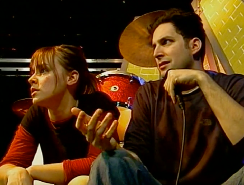Two cast members sitting in front of a drum kit on the set of Nate on Drums