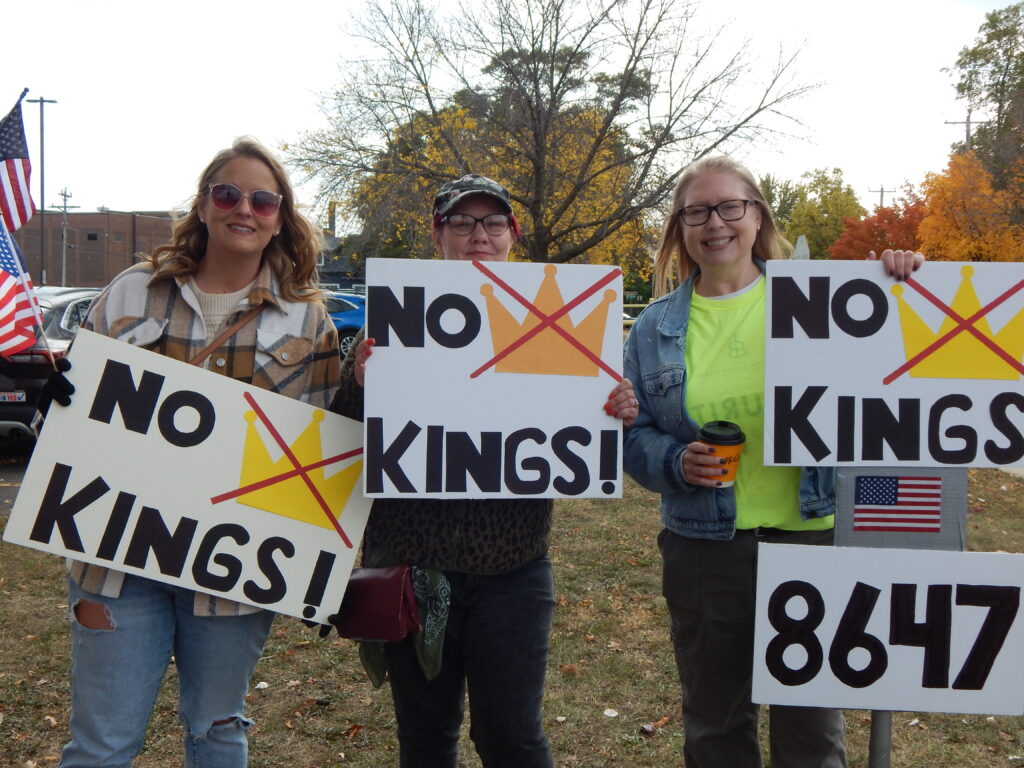 Three women stand together holding signs at the No Kings protest in St. Cloud