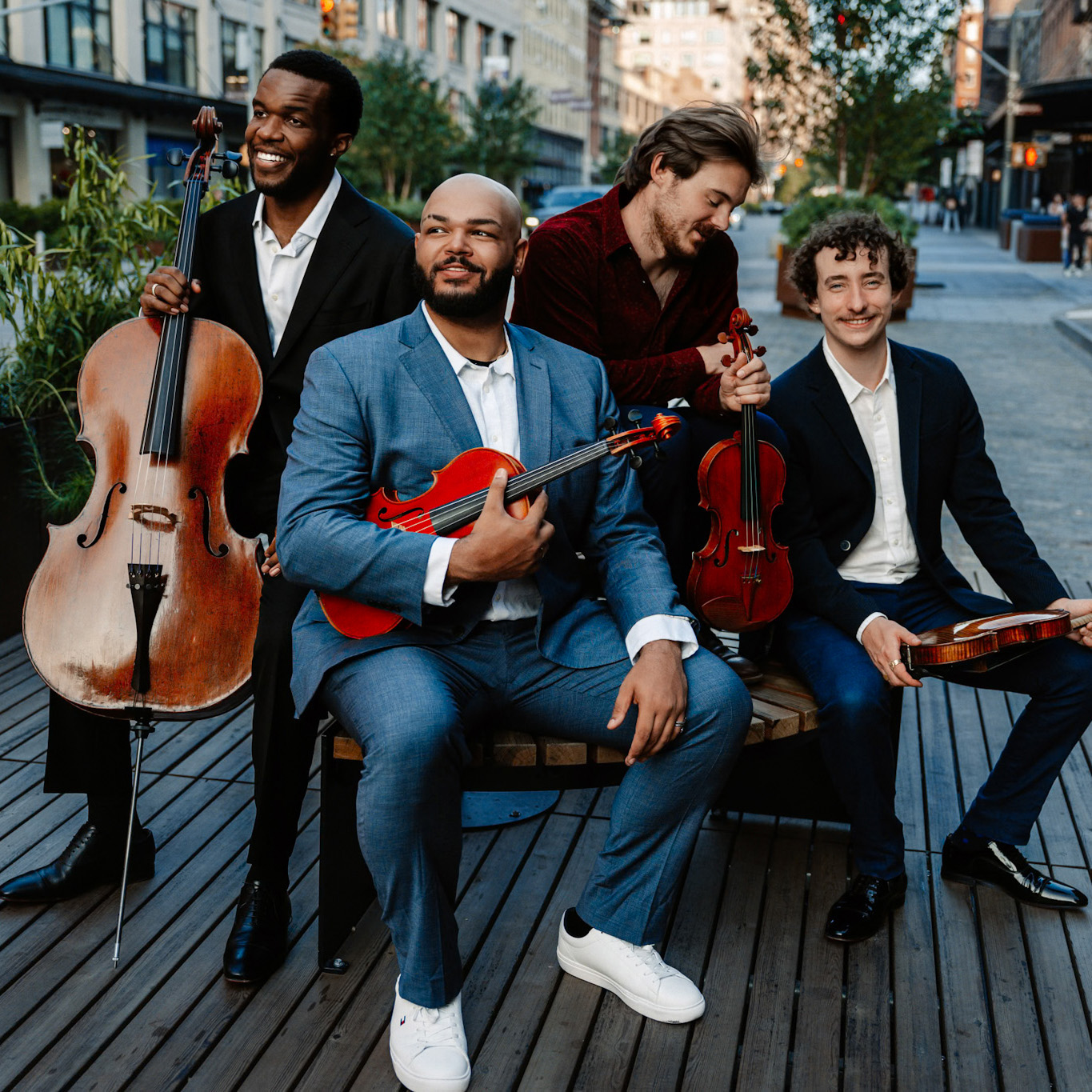 The members of the Isidore String Quartet sit with their instruments on a wooden platform near a city street