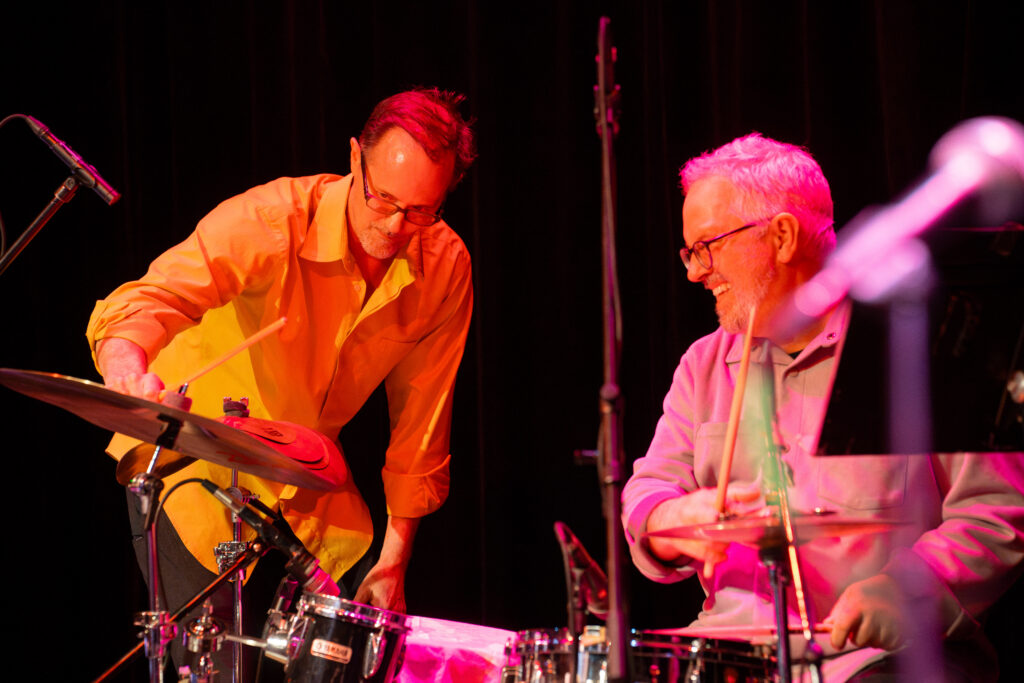 Matt Wilson taps a cymbal with a drumstick as the drummer looks on with a smile