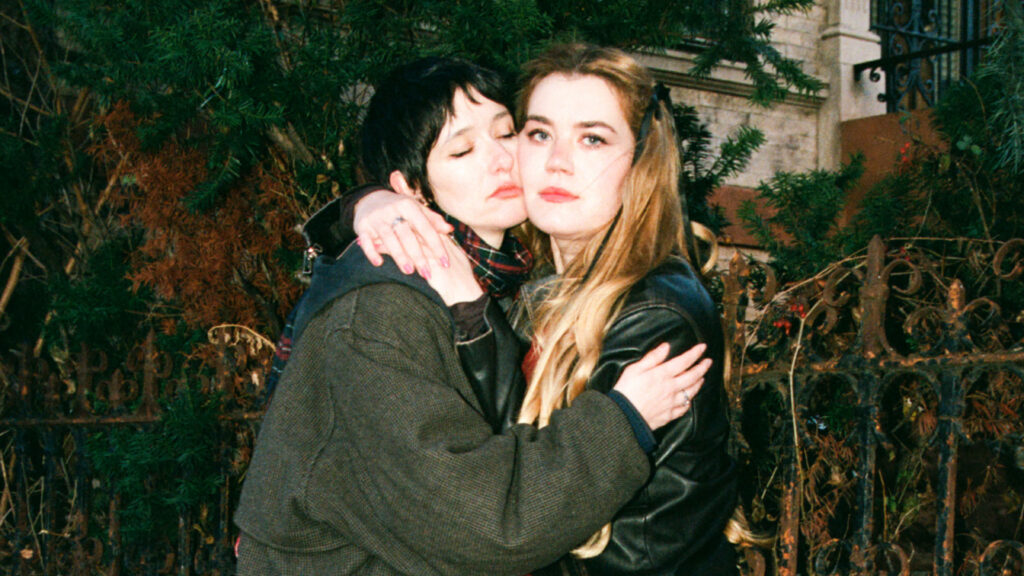 The two members of the band Daisy the Great stand holding each other, cheek to cheek