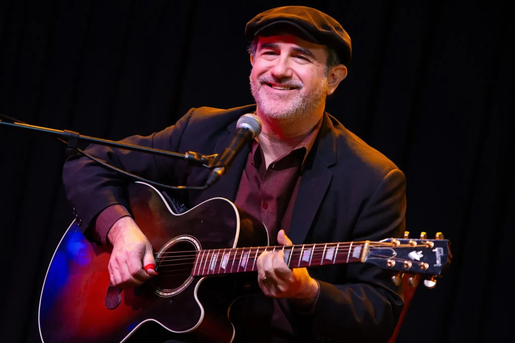 Jon Shain plays his guitar and smiles behind a microphone