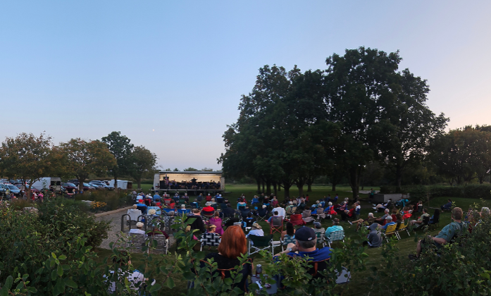 A crowd sits on a large lawn listening to a live music performance