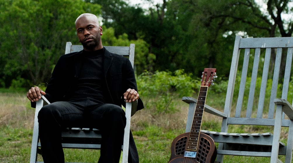 Buffalo Nichols sits in a chair. His guitar is leaned up against another chair that's next to him