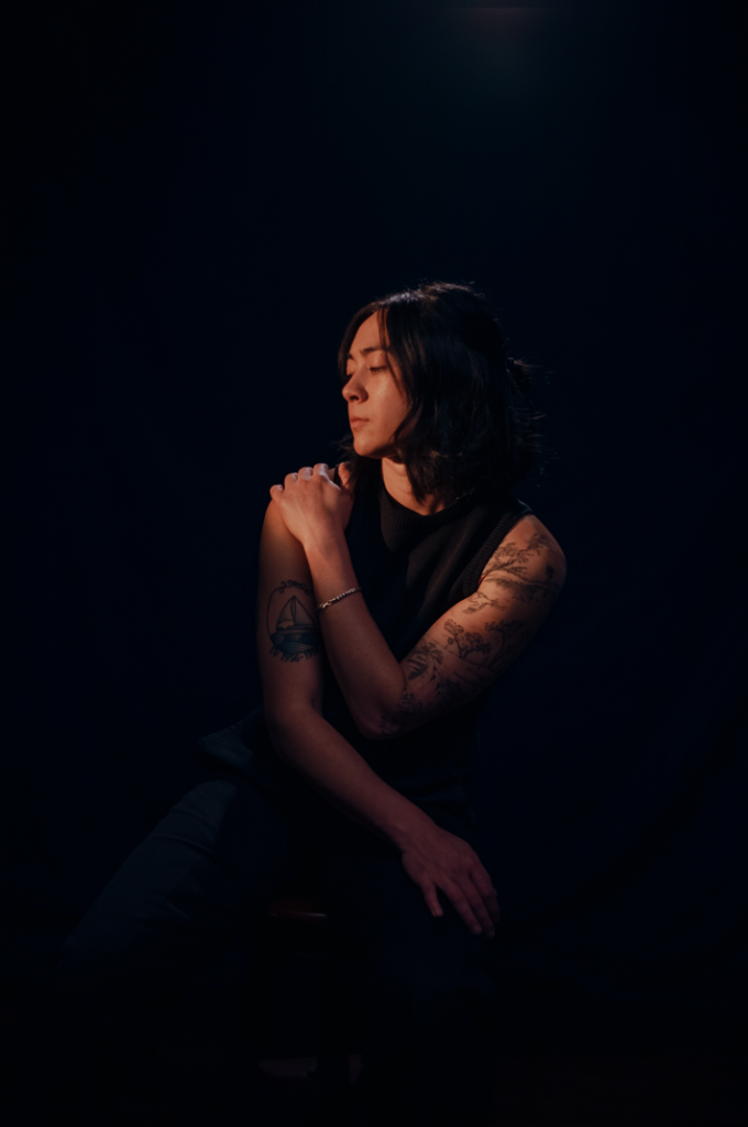 Lexi Vega, known as Mini Trees, sits on a stool and looks to the side. Photograph by Seannie Bryan