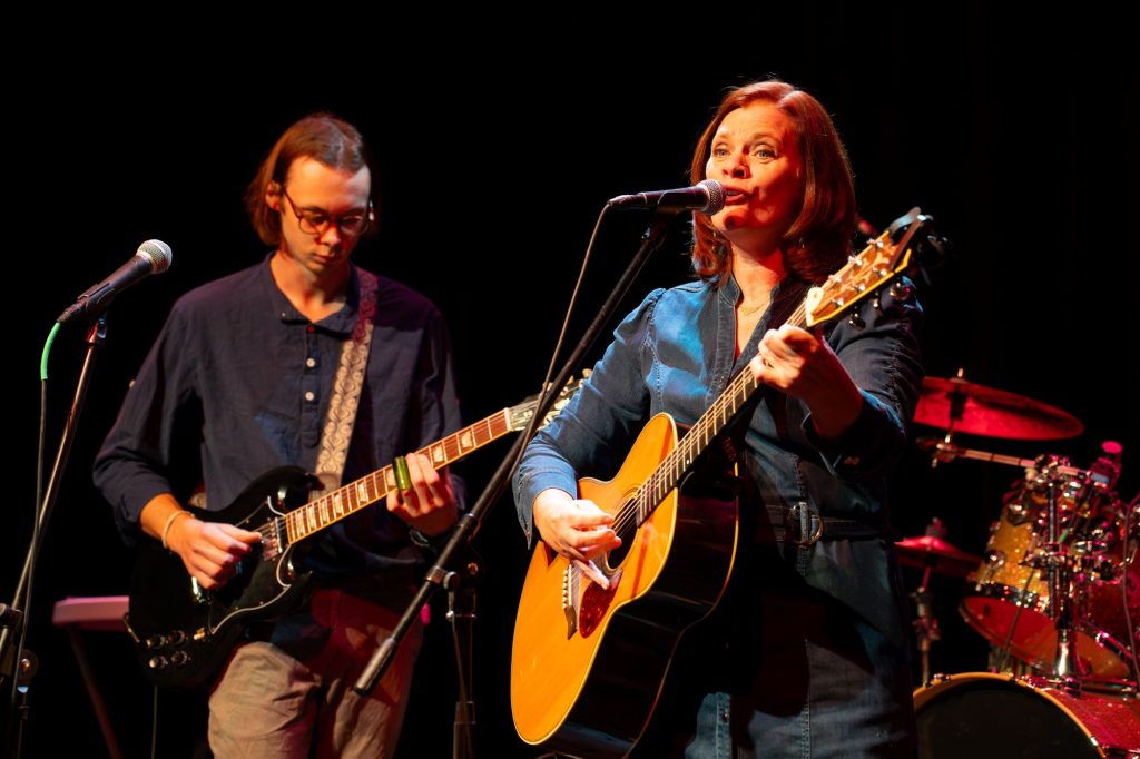 Katy Tessman performs on stage while her son plays guitar behind her